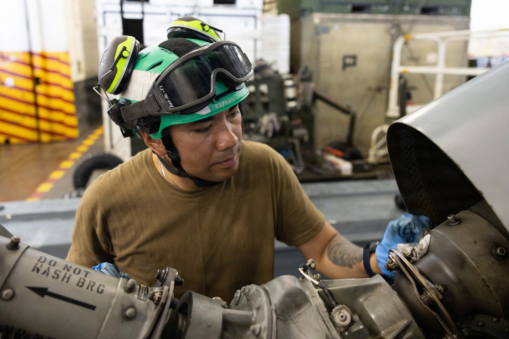 A military or aviation technician wearing a green helmet with goggles and blue nitrile gloves is performing maintenance on what appears to be an aircraft engine or mechanical assembly. The worker is focused on inspecting or servicing a component with visible labeling. The setting appears to be an aircraft hangar or flight deck aboard a naval vessel.