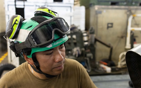 A military or aviation technician wearing a green helmet with goggles and blue nitrile gloves is performing maintenance on what appears to be an aircraft engine or mechanical assembly. The worker is focused on inspecting or servicing a component with visible labeling. The setting appears to be an aircraft hangar or flight deck aboard a naval vessel.