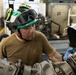 A military or aviation technician wearing a green helmet with goggles and blue nitrile gloves is performing maintenance on what appears to be an aircraft engine or mechanical assembly. The worker is focused on inspecting or servicing a component with visible labeling. The setting appears to be an aircraft hangar or flight deck aboard a naval vessel.