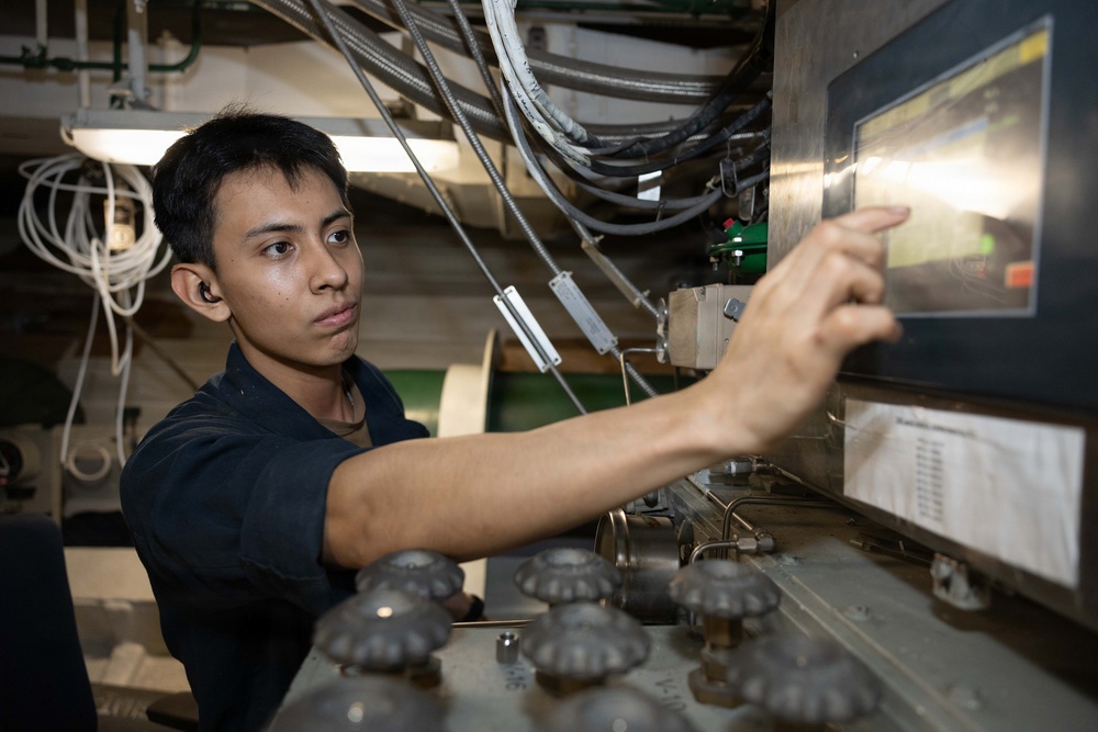 A young technician or military personnel in a dark uniform is operating or adjusting a touchscreen control panel in what appears to be an industrial or naval vessel environment. The workspace is densely populated with cables, conduits, and mechanical components. The individual appears focused and is interacting with a monitor displaying information.