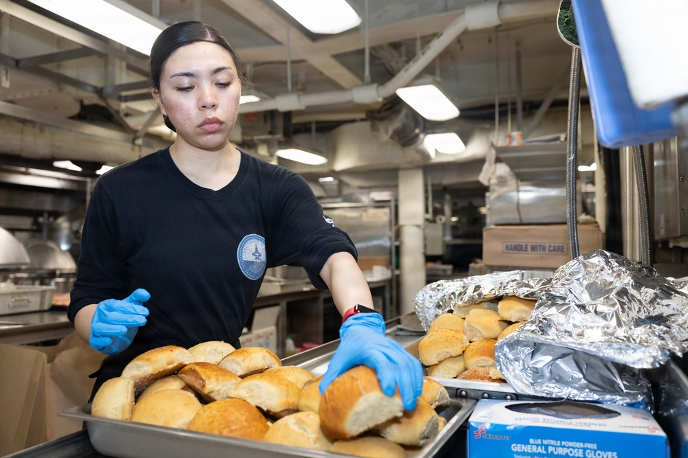 A food service worker wearing a black uniform with an insignia patch and blue nitrile gloves is handling dinner rolls in a large institutional kitchen. She is arranging or transferring bread rolls between metal sheet pans, with some rolls also covered in aluminum foil nearby. The setting appears to be a military or institutional galley/cafeteria kitchen based on the environment and uniform.