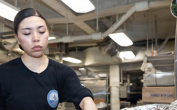 A food service worker wearing a black uniform with an insignia patch and blue nitrile gloves is handling dinner rolls in a large institutional kitchen. She is arranging or transferring bread rolls between metal sheet pans, with some rolls also covered in aluminum foil nearby. The setting appears to be a military or institutional galley/cafeteria kitchen based on the environment and uniform.