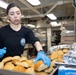 A food service worker wearing a black uniform with an insignia patch and blue nitrile gloves is handling dinner rolls in a large institutional kitchen. She is arranging or transferring bread rolls between metal sheet pans, with some rolls also covered in aluminum foil nearby. The setting appears to be a military or institutional galley/cafeteria kitchen based on the environment and uniform.
