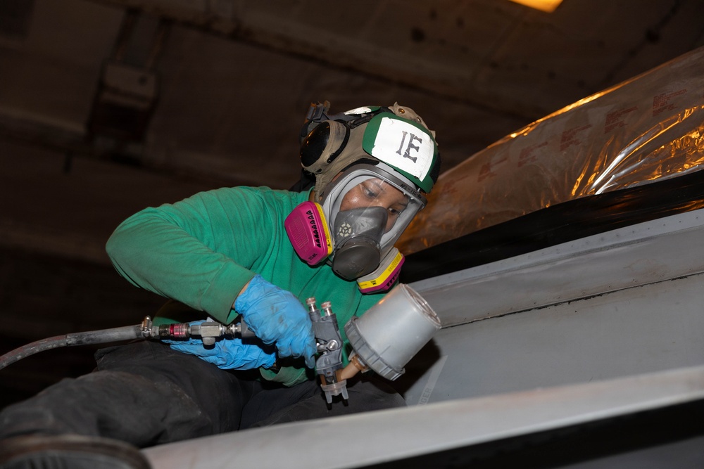 A worker wearing full respiratory protection and safety gear is using a spray paint gun to apply paint or coating to a surface, likely in an industrial or military vessel environment. The worker is equipped with a full-face respirator mask, green helmet marked 'IE', blue nitrile gloves, and a green long-sleeve shirt. The confined working space and protective equipment suggest this is a maintenance or refurbishment operation in a potentially hazardous environment.