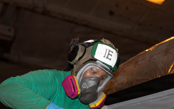 A worker wearing full respiratory protection and safety gear is using a spray paint gun to apply paint or coating to a surface, likely in an industrial or military vessel environment. The worker is equipped with a full-face respirator mask, green helmet marked 'IE', blue nitrile gloves, and a green long-sleeve shirt. The confined working space and protective equipment suggest this is a maintenance or refurbishment operation in a potentially hazardous environment.
