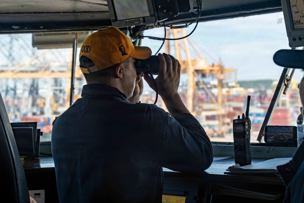 A naval officer wearing a yellow 'OOD' (Officer of the Deck) cap is looking through binoculars from what appears to be a ship's bridge or pilothouse. The setting overlooks a busy commercial port with large container cranes and stacked shipping containers visible in the background. The bridge is equipped with navigation and communication equipment.
