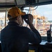 A naval officer wearing a yellow 'OOD' (Officer of the Deck) cap is looking through binoculars from what appears to be a ship's bridge or pilothouse. The setting overlooks a busy commercial port with large container cranes and stacked shipping containers visible in the background. The bridge is equipped with navigation and communication equipment.