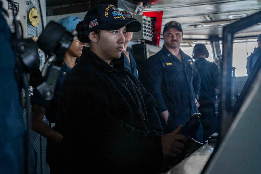The image shows U.S. Navy personnel on the bridge of a naval vessel, likely the USS Abraham Lincoln based on the cap insignia visible on the foreground sailor. A female sailor in the foreground appears to be at the helm or a control station, with several other crew members standing behind her. The setting is a ship's bridge or combat information center with various navigation and control equipment visible.