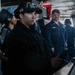 The image shows U.S. Navy personnel on the bridge of a naval vessel, likely the USS Abraham Lincoln based on the cap insignia visible on the foreground sailor. A female sailor in the foreground appears to be at the helm or a control station, with several other crew members standing behind her. The setting is a ship's bridge or combat information center with various navigation and control equipment visible.