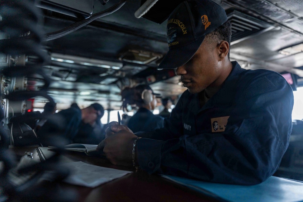A U.S. Navy sailor is writing in a logbook or document while stationed aboard what appears to be the USS Abraham Lincoln, as indicated by the text on their cap. The sailor is wearing standard Navy working uniform (NWU) and is focused on documentation tasks in what appears to be a ship's bridge or interior workspace. Several other personnel are visible in the background in a similar naval environment.