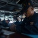 A U.S. Navy sailor is writing in a logbook or document while stationed aboard what appears to be the USS Abraham Lincoln, as indicated by the text on their cap. The sailor is wearing standard Navy working uniform (NWU) and is focused on documentation tasks in what appears to be a ship's bridge or interior workspace. Several other personnel are visible in the background in a similar naval environment.