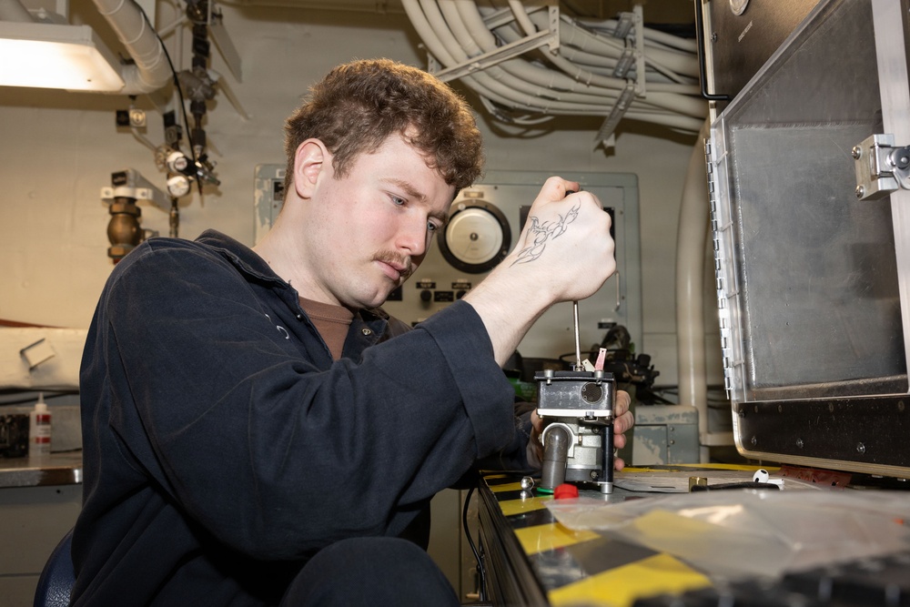 A young man with curly reddish-brown hair and a mustache is using a screwdriver to work on a mechanical component inside what appears to be a naval vessel or industrial facility. He is wearing a dark navy coverall uniform and has a visible tattoo on his right hand. The environment features overhead pipes, cable bundles, and mounted equipment consistent with a ship's interior workspace.