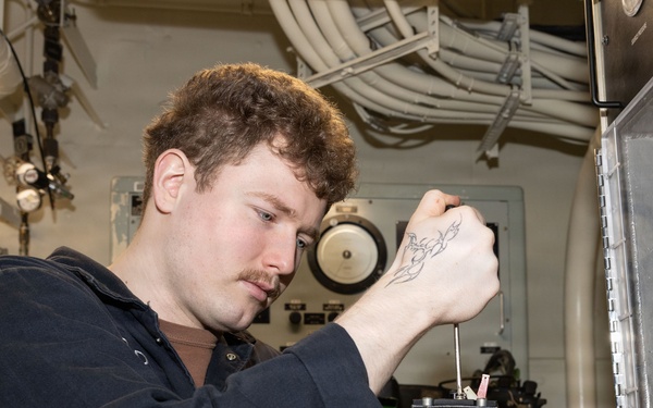 A young man with curly reddish-brown hair and a mustache is using a screwdriver to work on a mechanical component inside what appears to be a naval vessel or industrial facility. He is wearing a dark navy coverall uniform and has a visible tattoo on his right hand. The environment features overhead pipes, cable bundles, and mounted equipment consistent with a ship's interior workspace.