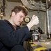 A young man with curly reddish-brown hair and a mustache is using a screwdriver to work on a mechanical component inside what appears to be a naval vessel or industrial facility. He is wearing a dark navy coverall uniform and has a visible tattoo on his right hand. The environment features overhead pipes, cable bundles, and mounted equipment consistent with a ship's interior workspace.
