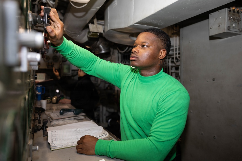 A young man wearing a green long-sleeve turtleneck shirt is working in what appears to be a military vessel or industrial control room. He is reaching upward to adjust or inspect equipment mounted overhead while standing at a workstation with open logbooks or documentation. A second person is visible in the background, also working at the same station.