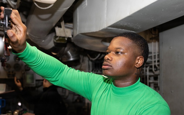 A young man wearing a green long-sleeve turtleneck shirt is working in what appears to be a military vessel or industrial control room. He is reaching upward to adjust or inspect equipment mounted overhead while standing at a workstation with open logbooks or documentation. A second person is visible in the background, also working at the same station.