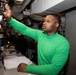 A young man wearing a green long-sleeve turtleneck shirt is working in what appears to be a military vessel or industrial control room. He is reaching upward to adjust or inspect equipment mounted overhead while standing at a workstation with open logbooks or documentation. A second person is visible in the background, also working at the same station.