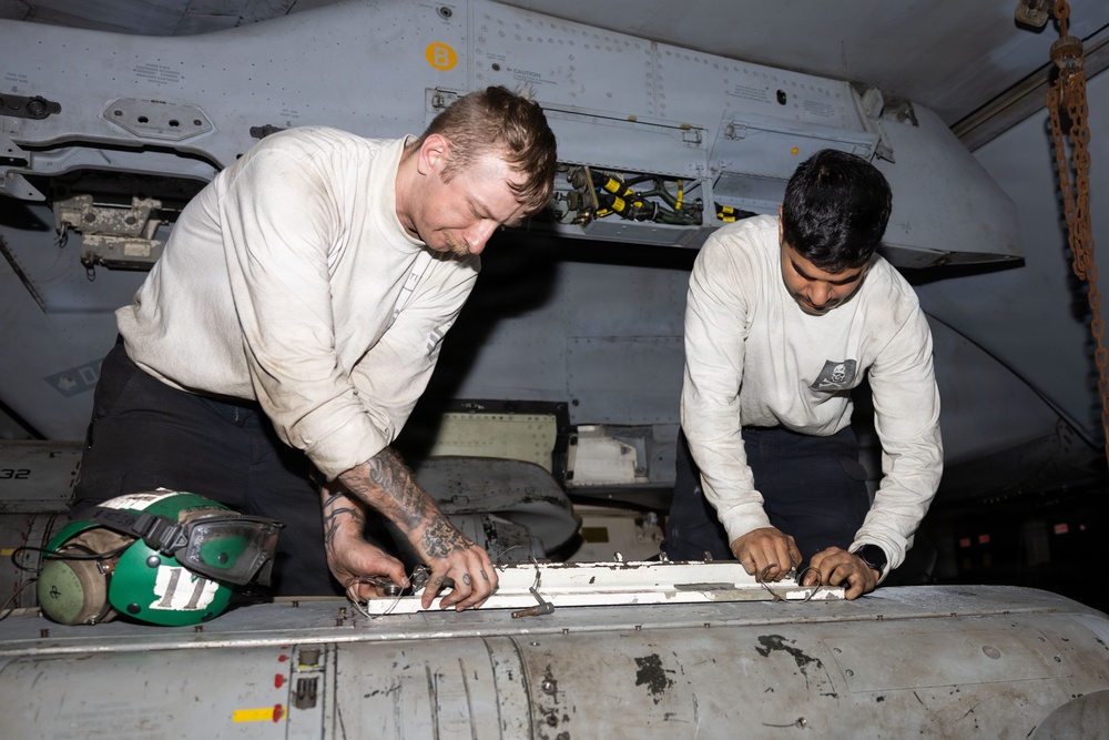 Two military aviation maintenance personnel in white long-sleeve shirts are working on an aircraft component, appearing to perform maintenance or repair on a panel or access door. Both individuals are leaning over the work surface in what appears to be an aircraft hangar bay or below-deck maintenance area. A green flight deck cranial helmet is visible nearby, indicating a naval aviation environment.