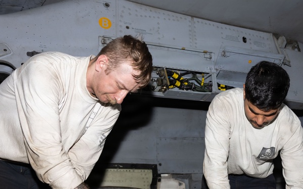 Two military aviation maintenance personnel in white long-sleeve shirts are working on an aircraft component, appearing to perform maintenance or repair on a panel or access door. Both individuals are leaning over the work surface in what appears to be an aircraft hangar bay or below-deck maintenance area. A green flight deck cranial helmet is visible nearby, indicating a naval aviation environment.