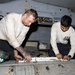 Two military aviation maintenance personnel in white long-sleeve shirts are working on an aircraft component, appearing to perform maintenance or repair on a panel or access door. Both individuals are leaning over the work surface in what appears to be an aircraft hangar bay or below-deck maintenance area. A green flight deck cranial helmet is visible nearby, indicating a naval aviation environment.