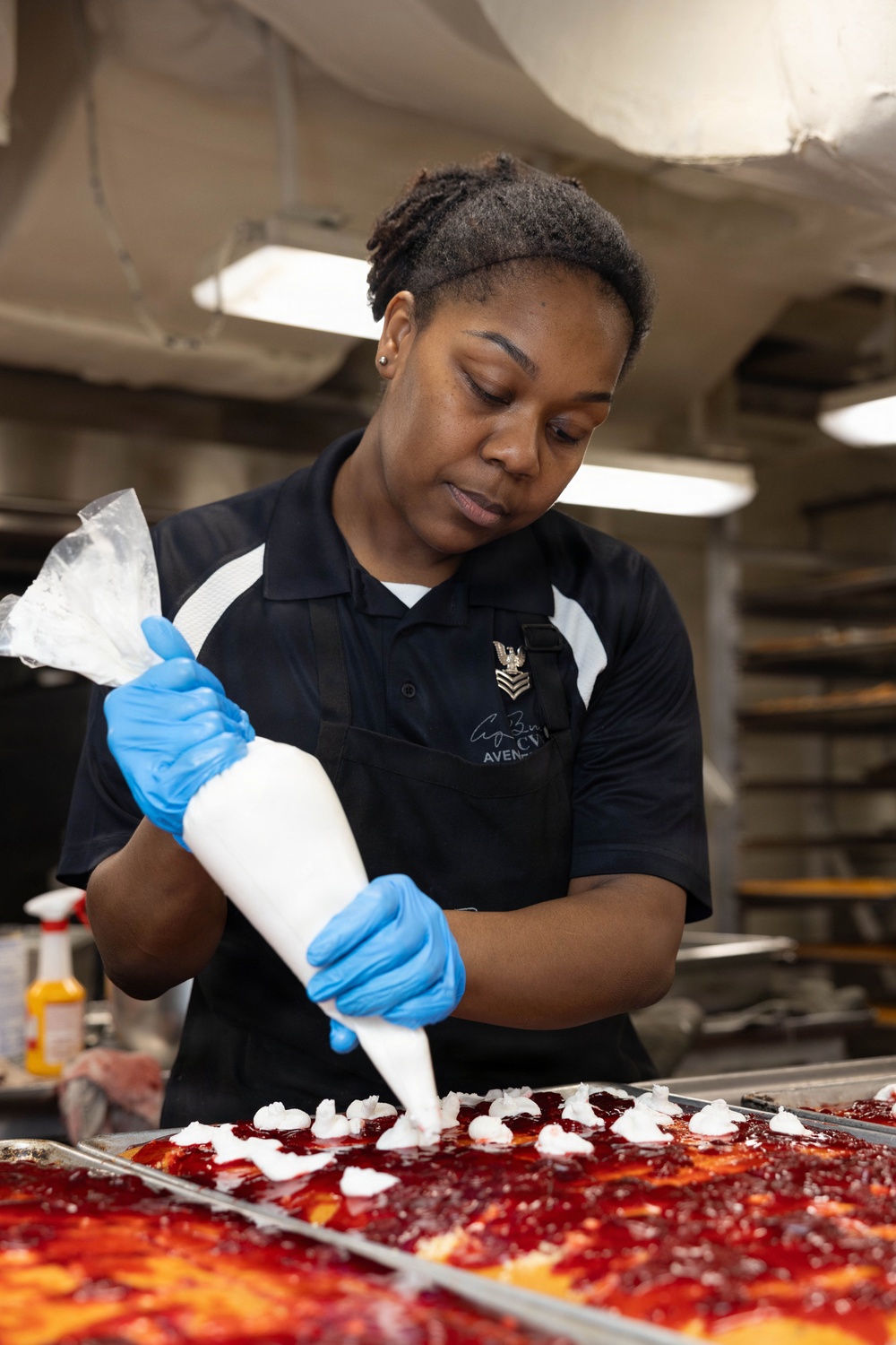 A U.S. Navy culinary specialist, identifiable by a Petty Officer rank insignia pin on her shirt, is piping white cream or whipped topping onto sheet pans of desserts topped with red fruit filling. She is working in what appears to be a military galley kitchen, wearing blue nitrile gloves and a black apron. The setting suggests large-scale food preparation aboard a naval vessel.