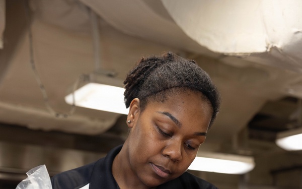 A U.S. Navy culinary specialist, identifiable by a Petty Officer rank insignia pin on her shirt, is piping white cream or whipped topping onto sheet pans of desserts topped with red fruit filling. She is working in what appears to be a military galley kitchen, wearing blue nitrile gloves and a black apron. The setting suggests large-scale food preparation aboard a naval vessel.