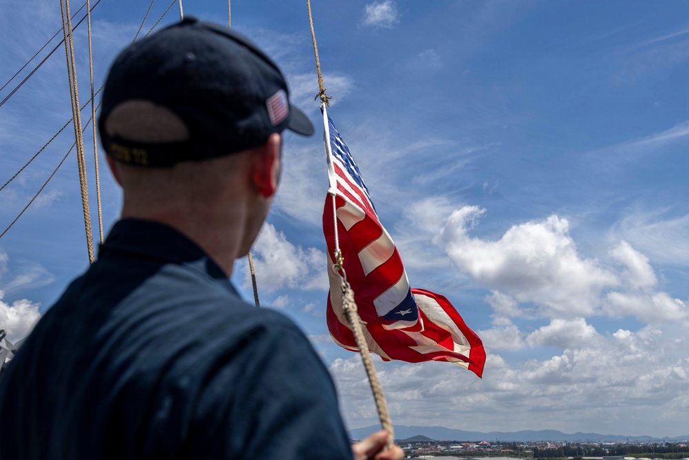A U.S. Navy sailor is seen from behind on the deck of a naval vessel, holding a rope attached to a flagpole flying the American flag. The flag appears to be in motion due to wind, and the scene takes place under a partly cloudy sky with a coastal cityscape visible in the background. The sailor is wearing a dark Navy uniform and a cap with a small American flag patch and visible text.