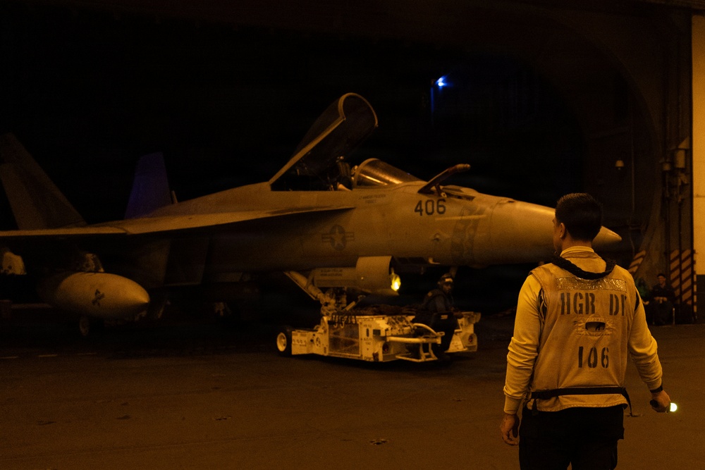 A nighttime scene in what appears to be an aircraft carrier hangar bay, showing a U.S. Navy F/A-18 Hornet or Super Hornet with tail number 406 being moved or serviced. A crew member wearing a vest marked 'HGR DK 106' (Hangar Deck 106) stands observing the aircraft. Ground support equipment, including a tow vehicle, is visible near the aircraft.