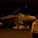 A nighttime scene in what appears to be an aircraft carrier hangar bay, showing a U.S. Navy F/A-18 Hornet or Super Hornet with tail number 406 being moved or serviced. A crew member wearing a vest marked 'HGR DK 106' (Hangar Deck 106) stands observing the aircraft. Ground support equipment, including a tow vehicle, is visible near the aircraft.