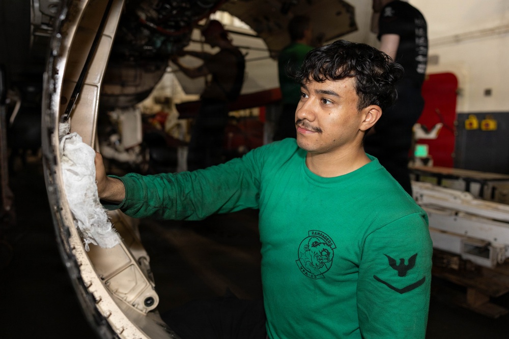 A U.S. Navy sailor wearing a green long-sleeve shirt with a 'Rampagers VFA-83' squadron patch is performing maintenance on what appears to be a military aircraft in a hangar bay. He is wiping or cleaning a circular aircraft component, likely an engine intake or landing gear housing. Additional personnel are visible working in the background.