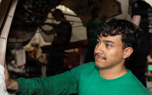 A U.S. Navy sailor wearing a green long-sleeve shirt with a 'Rampagers VFA-83' squadron patch is performing maintenance on what appears to be a military aircraft in a hangar bay. He is wiping or cleaning a circular aircraft component, likely an engine intake or landing gear housing. Additional personnel are visible working in the background.