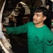 A U.S. Navy sailor wearing a green long-sleeve shirt with a 'Rampagers VFA-83' squadron patch is performing maintenance on what appears to be a military aircraft in a hangar bay. He is wiping or cleaning a circular aircraft component, likely an engine intake or landing gear housing. Additional personnel are visible working in the background.