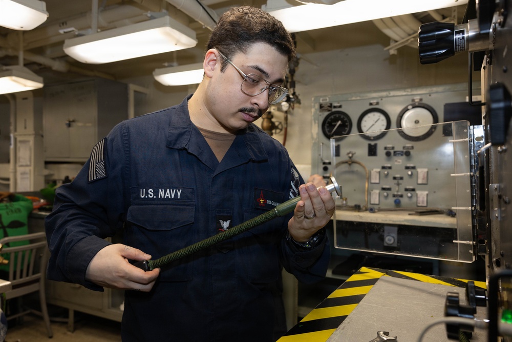 A U.S. Navy sailor in Navy Working Uniform (NWU) is inspecting or handling a cylindrical tool or component in what appears to be a shipboard workspace or machinery room. The sailor wears glasses and has a name tag and rank insignia visible on their uniform. The background shows gauges, control panels, and industrial equipment consistent with a naval vessel interior.