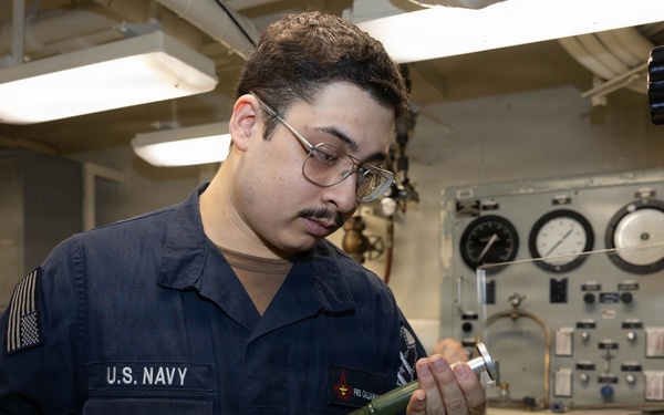 A U.S. Navy sailor in Navy Working Uniform (NWU) is inspecting or handling a cylindrical tool or component in what appears to be a shipboard workspace or machinery room. The sailor wears glasses and has a name tag and rank insignia visible on their uniform. The background shows gauges, control panels, and industrial equipment consistent with a naval vessel interior.