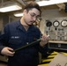 A U.S. Navy sailor in Navy Working Uniform (NWU) is inspecting or handling a cylindrical tool or component in what appears to be a shipboard workspace or machinery room. The sailor wears glasses and has a name tag and rank insignia visible on their uniform. The background shows gauges, control panels, and industrial equipment consistent with a naval vessel interior.