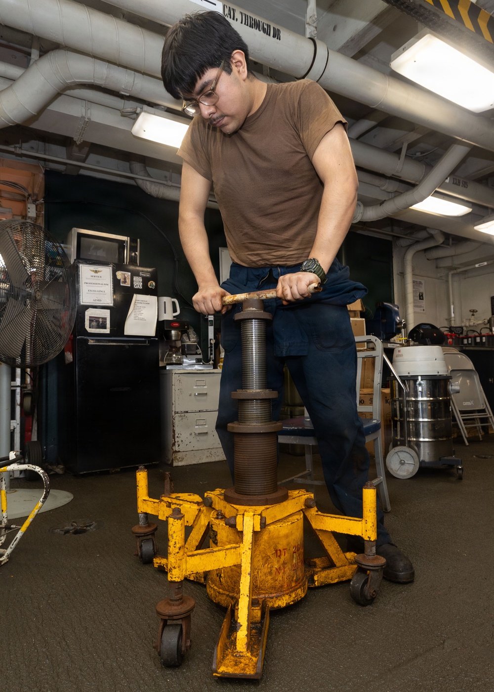 A young man wearing glasses and a brown t-shirt is operating a large yellow mechanical device with a threaded screw/jack mechanism in what appears to be a military ship's workshop or maintenance bay. He is applying force to a handle bar to turn the central threaded spindle of the yellow-painted equipment. The workspace contains typical shop equipment including a fan, filing cabinet, vacuum, and overhead piping.