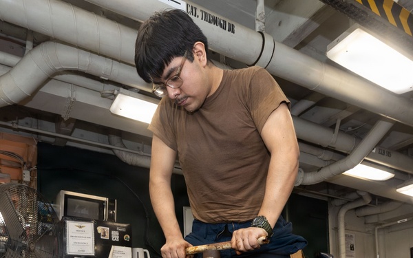 A young man wearing glasses and a brown t-shirt is operating a large yellow mechanical device with a threaded screw/jack mechanism in what appears to be a military ship's workshop or maintenance bay. He is applying force to a handle bar to turn the central threaded spindle of the yellow-painted equipment. The workspace contains typical shop equipment including a fan, filing cabinet, vacuum, and overhead piping.