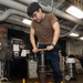 A young man wearing glasses and a brown t-shirt is operating a large yellow mechanical device with a threaded screw/jack mechanism in what appears to be a military ship's workshop or maintenance bay. He is applying force to a handle bar to turn the central threaded spindle of the yellow-painted equipment. The workspace contains typical shop equipment including a fan, filing cabinet, vacuum, and overhead piping.