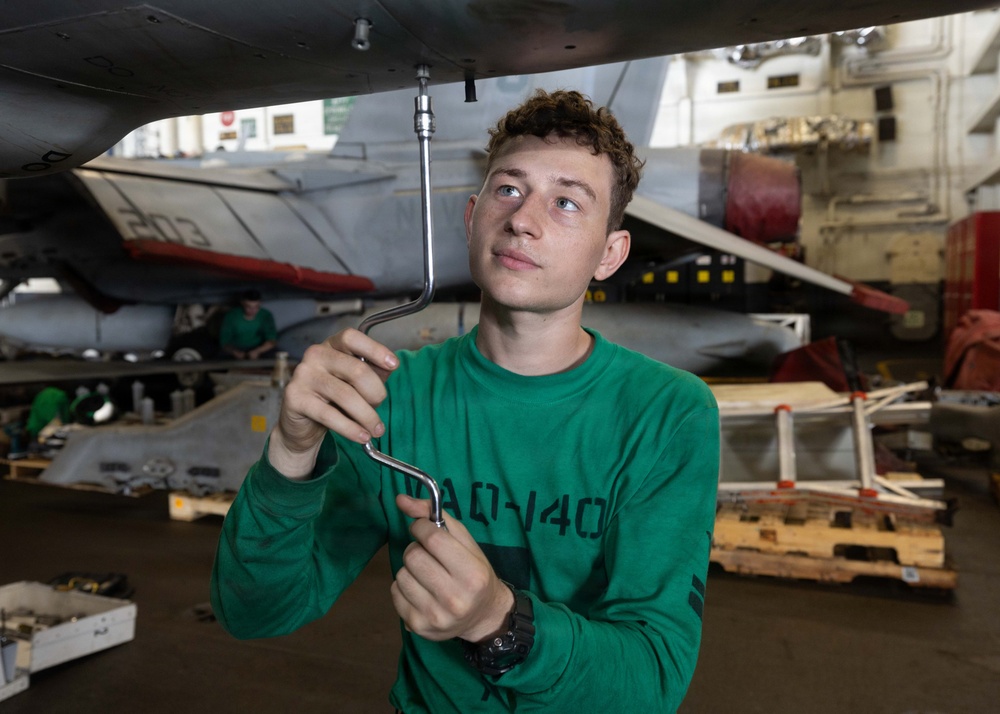 A young military technician in a green sweatshirt is performing maintenance work on the underside of a military aircraft, likely in a carrier hangar bay. He is using an L-shaped wrench or ratchet extension to work on a fitting or fastener on the aircraft's fuselage. The setting appears to be a naval vessel's hangar deck with multiple aircraft and equipment visible in the background.