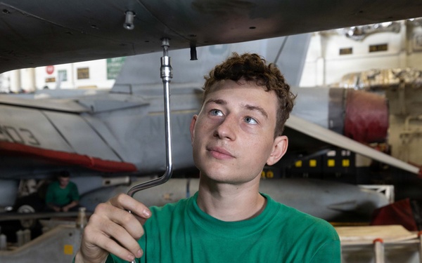 A young military technician in a green sweatshirt is performing maintenance work on the underside of a military aircraft, likely in a carrier hangar bay. He is using an L-shaped wrench or ratchet extension to work on a fitting or fastener on the aircraft's fuselage. The setting appears to be a naval vessel's hangar deck with multiple aircraft and equipment visible in the background.