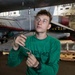 A young military technician in a green sweatshirt is performing maintenance work on the underside of a military aircraft, likely in a carrier hangar bay. He is using an L-shaped wrench or ratchet extension to work on a fitting or fastener on the aircraft's fuselage. The setting appears to be a naval vessel's hangar deck with multiple aircraft and equipment visible in the background.