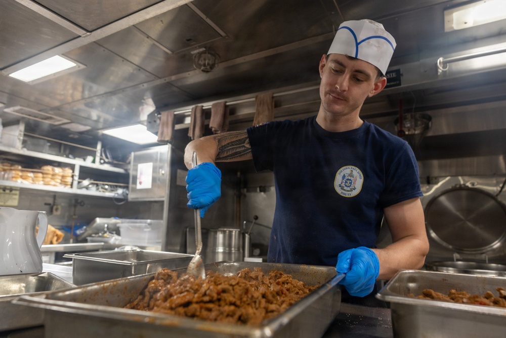 A young male cook or culinary specialist is working in a military ship's galley kitchen, stirring or serving a cooked meat dish from a large stainless steel pan. He is wearing a navy blue t-shirt with a ship's insignia, a white cook's hat with blue trim, and blue nitrile gloves. The stainless steel industrial kitchen environment and uniform suggest this is aboard a U.S. Navy vessel.