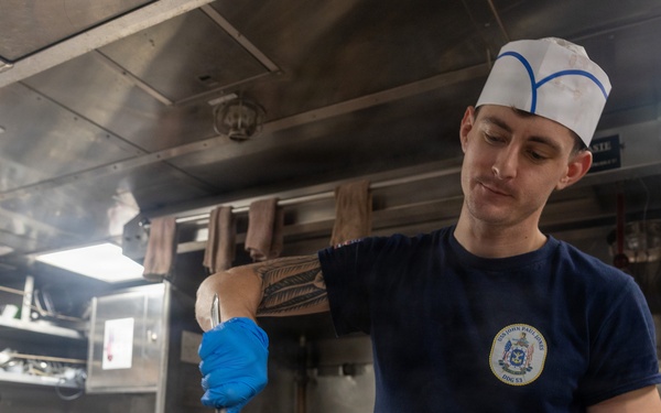 A young male cook or culinary specialist is working in a military ship's galley kitchen, stirring or serving a cooked meat dish from a large stainless steel pan. He is wearing a navy blue t-shirt with a ship's insignia, a white cook's hat with blue trim, and blue nitrile gloves. The stainless steel industrial kitchen environment and uniform suggest this is aboard a U.S. Navy vessel.