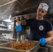 A young male cook or culinary specialist is working in a military ship's galley kitchen, stirring or serving a cooked meat dish from a large stainless steel pan. He is wearing a navy blue t-shirt with a ship's insignia, a white cook's hat with blue trim, and blue nitrile gloves. The stainless steel industrial kitchen environment and uniform suggest this is aboard a U.S. Navy vessel.