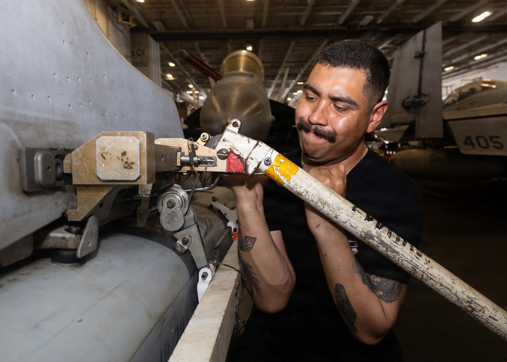 A military technician in a black t-shirt is performing maintenance work on an aircraft weapon or ordnance mounting system inside what appears to be a naval vessel's hangar bay. The worker is closely inspecting and manipulating a mechanical component attached to the underside or side of an aircraft. Additional aircraft and equipment are visible in the background of the enclosed hangar space.