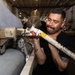 A military technician in a black t-shirt is performing maintenance work on an aircraft weapon or ordnance mounting system inside what appears to be a naval vessel's hangar bay. The worker is closely inspecting and manipulating a mechanical component attached to the underside or side of an aircraft. Additional aircraft and equipment are visible in the background of the enclosed hangar space.