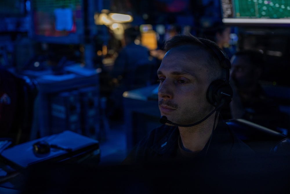 A man wearing a headset with a boom microphone is seated at a workstation in a dimly lit operations or command center environment. The room is bathed in blue ambient lighting with multiple screens and equipment visible in the background. His focused gaze suggests active monitoring or communication duties.