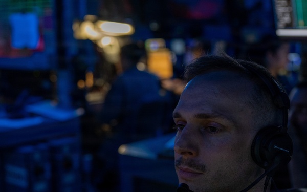 A man wearing a headset with a boom microphone is seated at a workstation in a dimly lit operations or command center environment. The room is bathed in blue ambient lighting with multiple screens and equipment visible in the background. His focused gaze suggests active monitoring or communication duties.