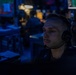 A man wearing a headset with a boom microphone is seated at a workstation in a dimly lit operations or command center environment. The room is bathed in blue ambient lighting with multiple screens and equipment visible in the background. His focused gaze suggests active monitoring or communication duties.