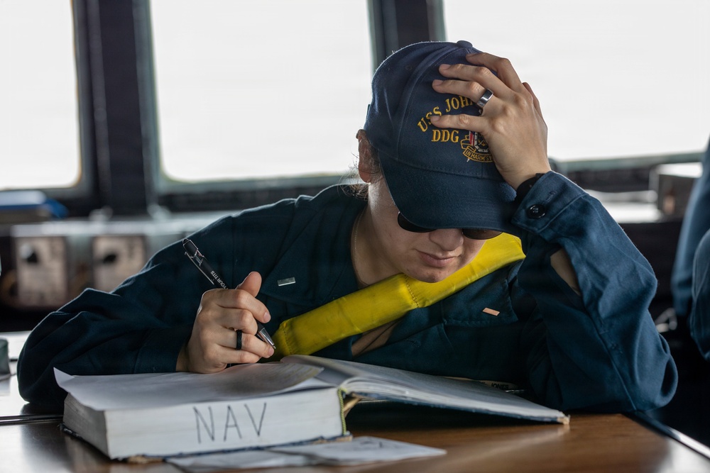 A U.S. Navy sailor in dark blue coveralls is seated at a desk aboard a naval vessel, writing in or reviewing a binder labeled 'NAV' (Navigation). The sailor is wearing a cap bearing 'USS John...' and 'DDG' designation, along with sunglasses and a yellow inflatable life vest/collar. The setting appears to be the bridge or a navigation workspace of a Navy destroyer.